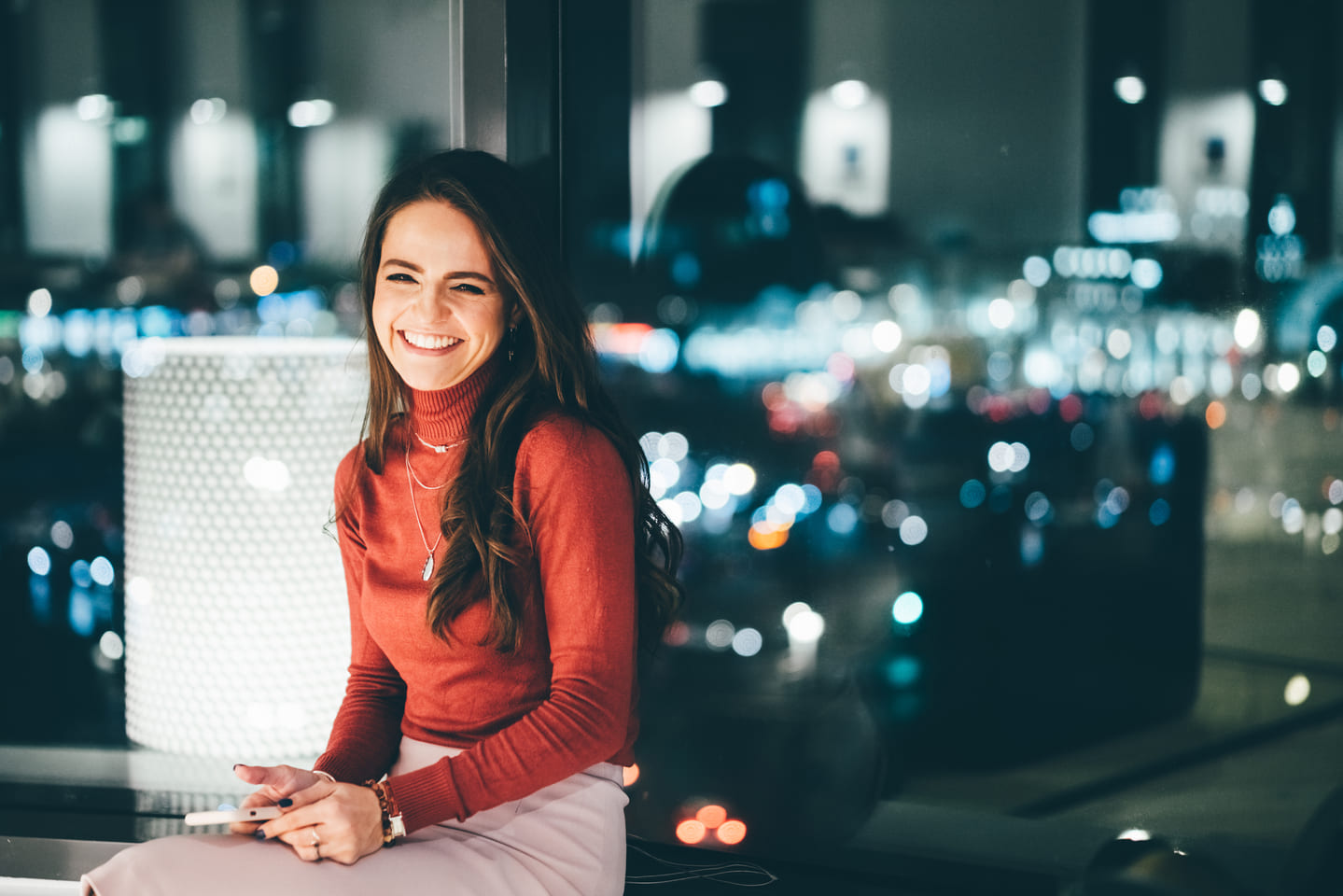 Portrait of a young woman, leaning against a window, city lights behind her, and holding a phone