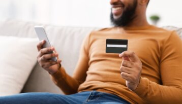Plan rapproché d'un homme noir, barbu, pull orange, tenant un téléphone dans une main et une carte bancaire dans l'autre